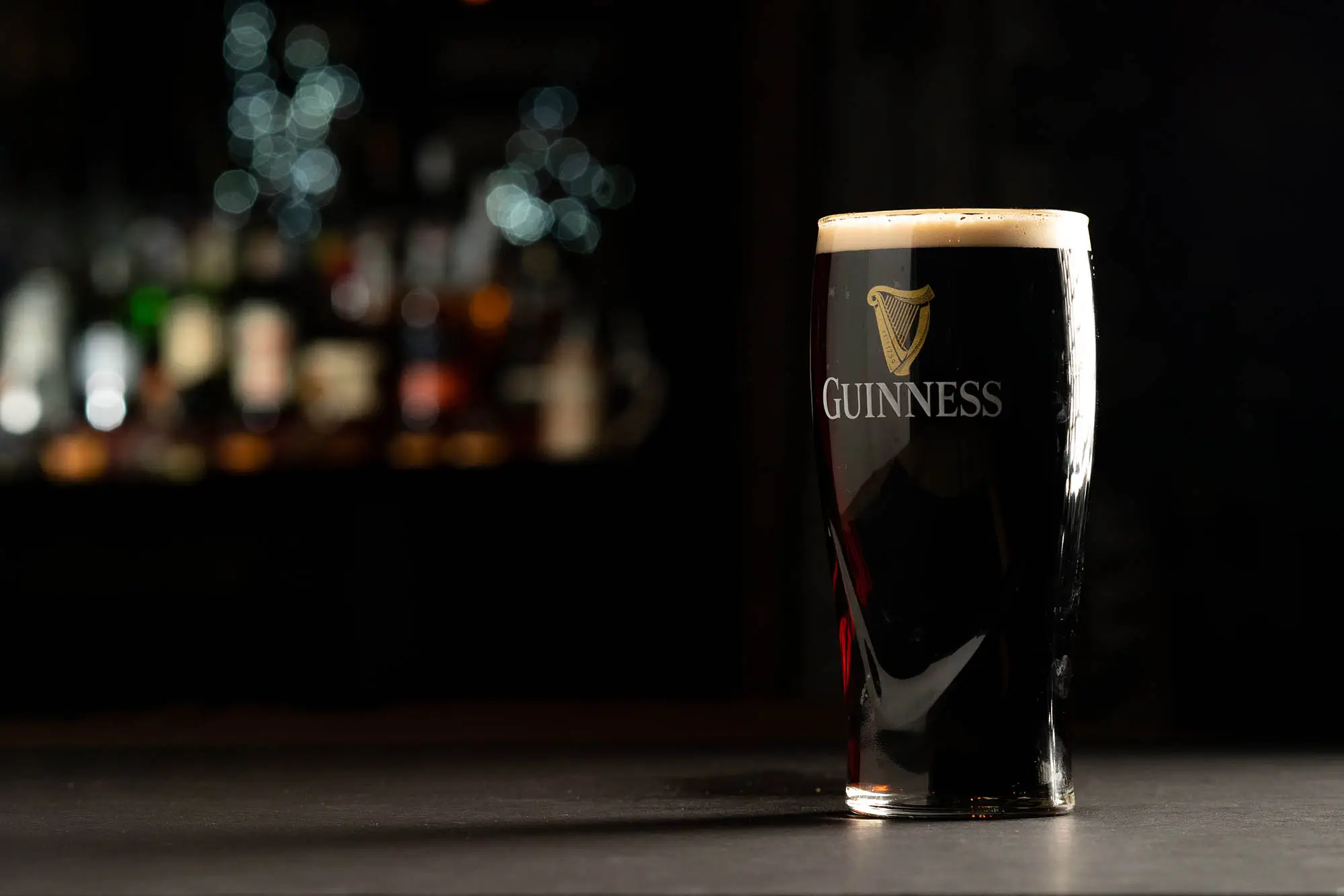 A pint of dark Guinness beer with foam head on a bar counter with blurred colorful bottles in the background.