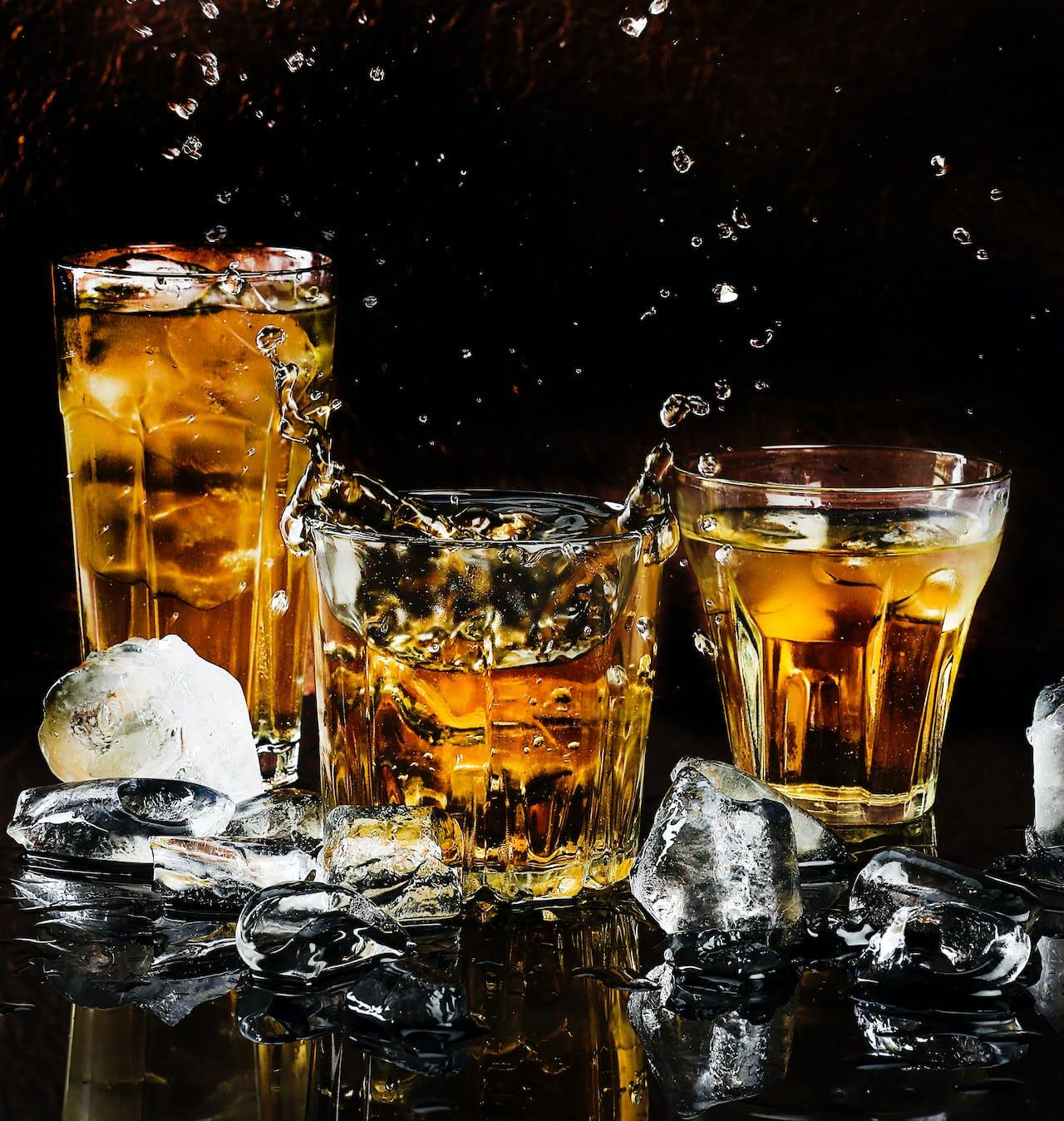 Three glasses of whiskey with ice cubes, one glass with a splash of liquid, surrounded by scattered ice cubes on a reflective surface.