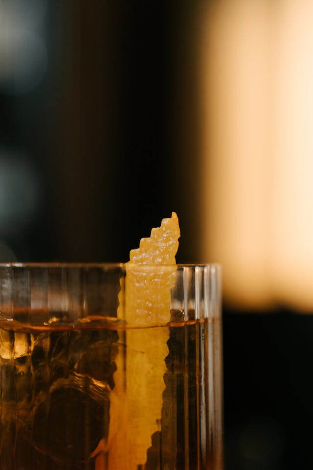 Glass with amber liquid and ice cubes garnished with a zigzag-edged lemon peel.