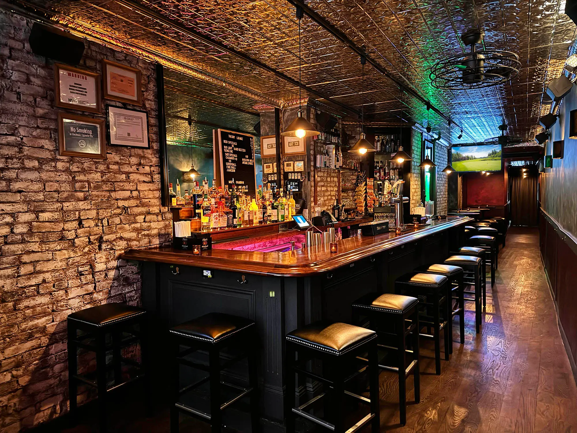 Interior of a long, narrow bar with a polished wooden counter, black bar stools, exposed brick walls, and a metallic patterned ceiling.