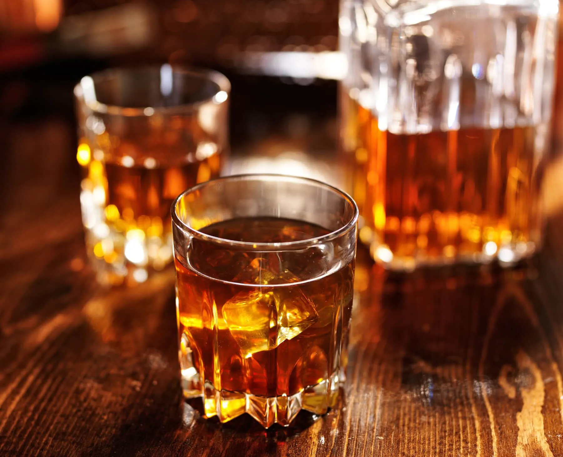 Glass of whiskey with ice on a wooden table with two additional glasses and a whiskey decanter in the background.