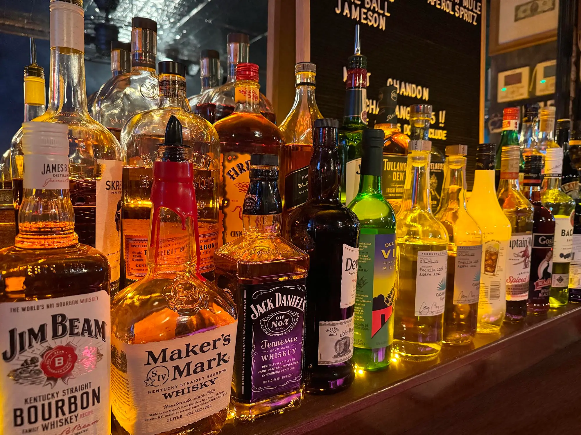 Assorted bottles of whiskey, bourbon, tequila, and other liquors lined up on a bar counter with a drink menu board in the background.