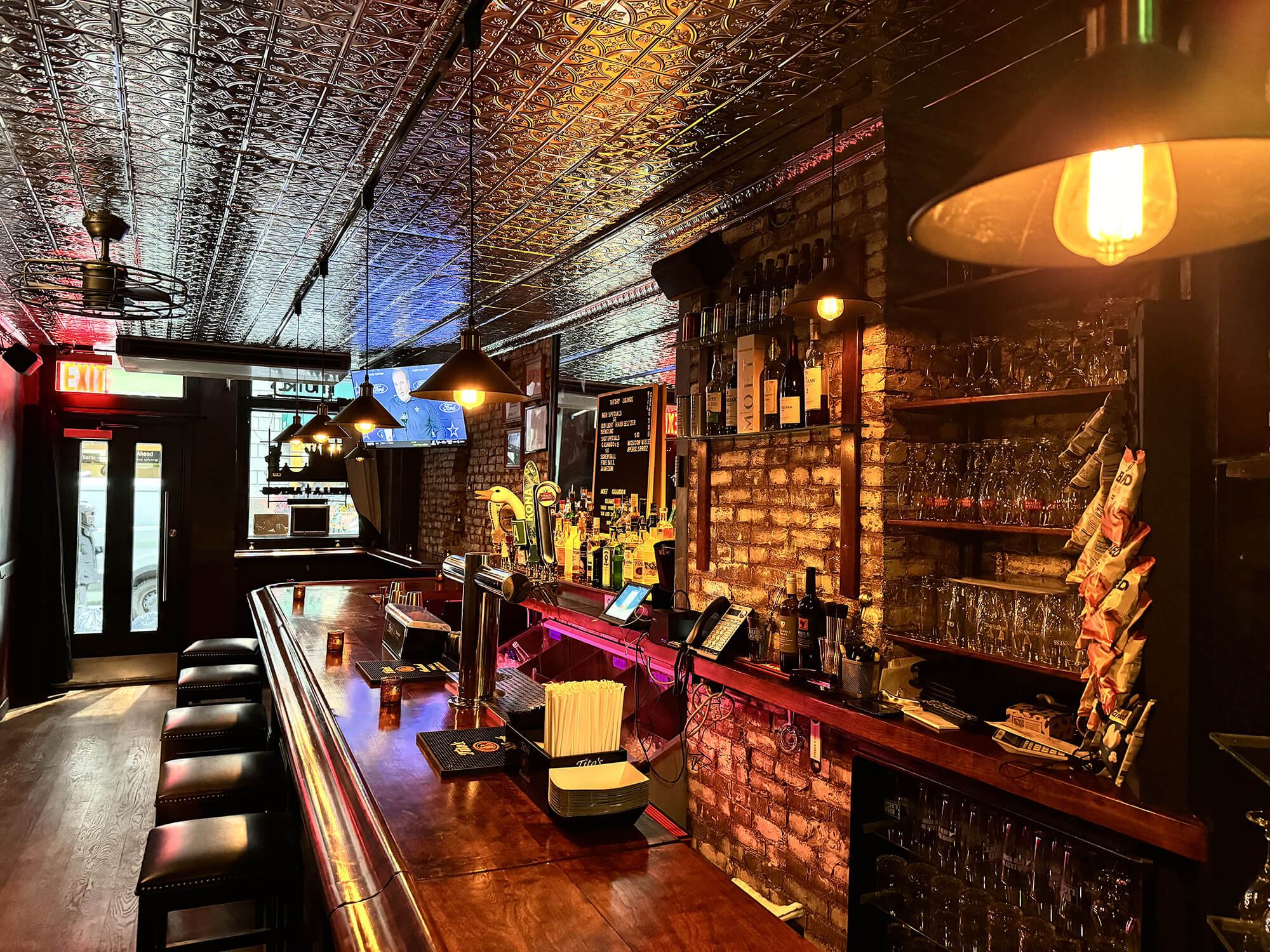 Cozy bar interior with wooden counter, leather stools, hanging pendant lights, shelves with bottles and glasses, and TV screens.