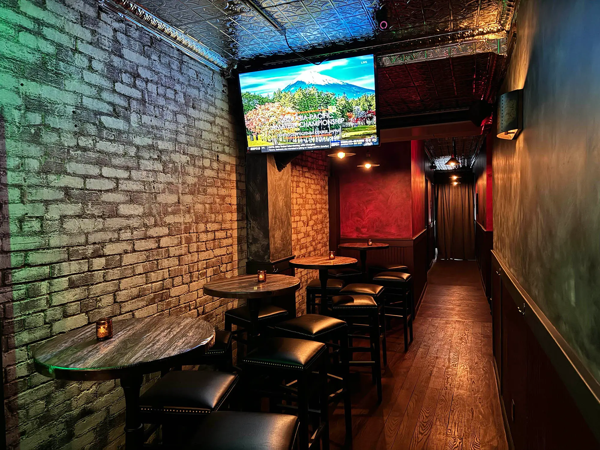 Dimly lit narrow bar area with high tables, black cushioned stools, exposed brick wall, and a TV screen displaying a golf championship.