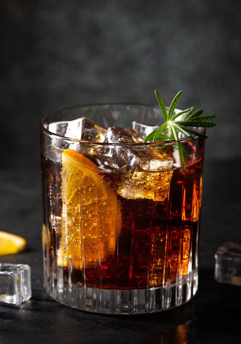 Glass of dark soda with ice cubes, a lemon slice, and a rosemary sprig garnish on a dark surface.
