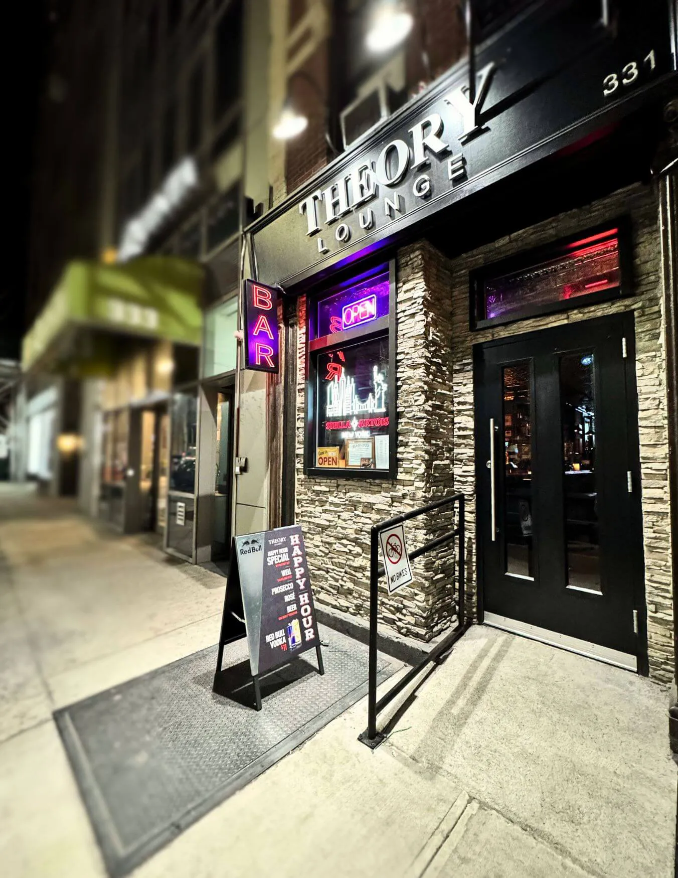 Night view of Theory Lounge bar entrance with neon 'BAR' and 'OPEN' signs, a happy hour chalkboard sign, and a no bikes sign on the railing.