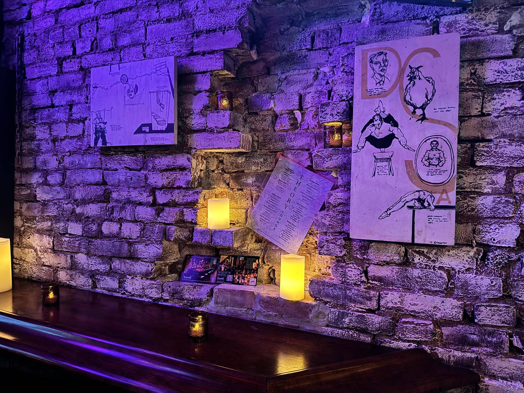 Exposed brick wall with purple lighting, decorated with sketches, a posted menu, and candles on a wooden surface.