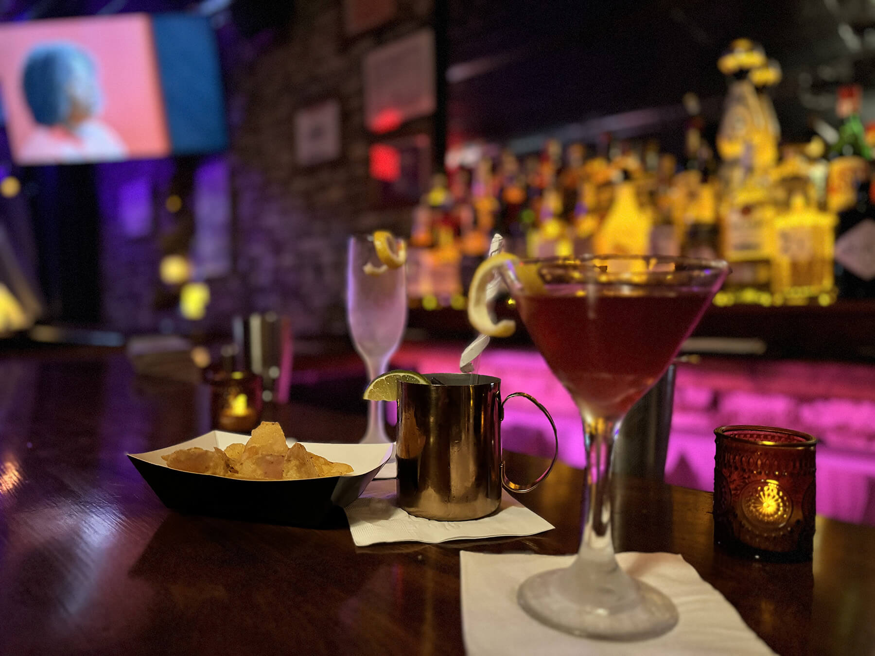 Close-up of cocktails and a bowl of chips on a bar counter with colorful bottles and a TV in the blurred background.
