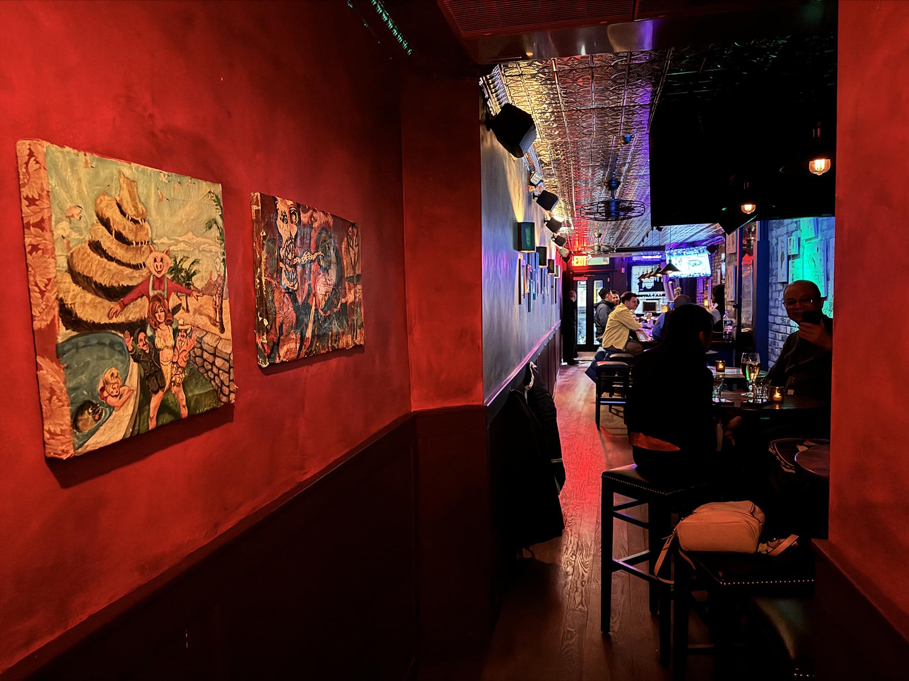 Dimly lit bar interior with red walls, two vibrant paintings on the left, and patrons sitting at tables and the bar.