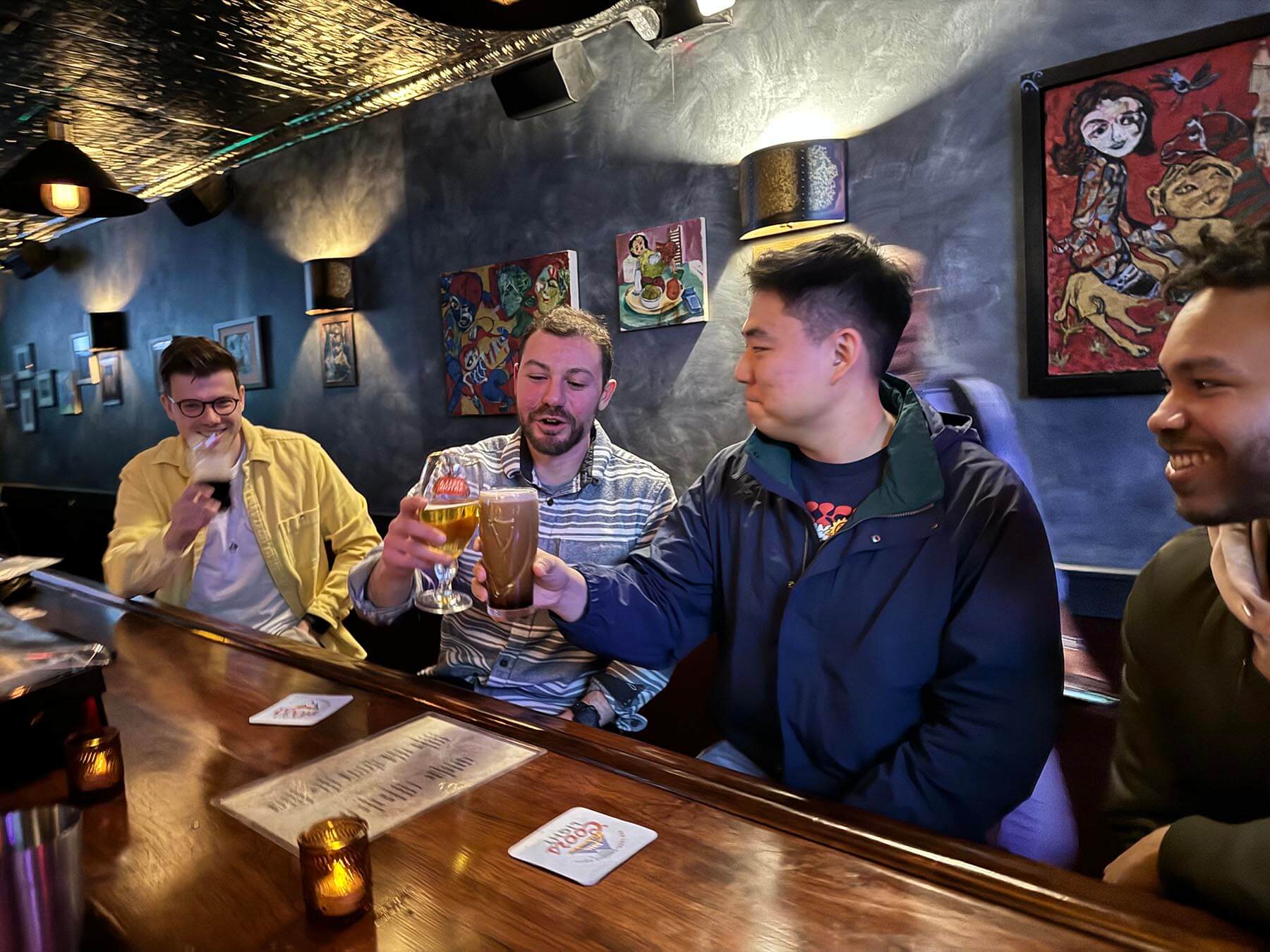 Four friends sitting at a bar, smiling and clinking their glasses of beer together.