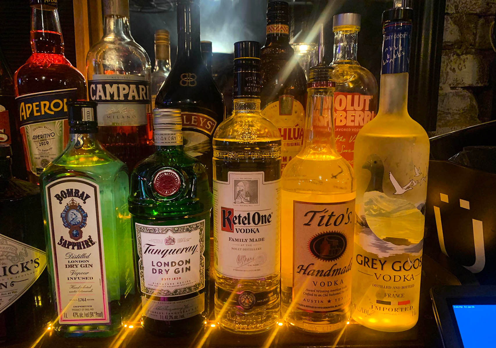 Various liquor bottles including Bombay Sapphire gin, Tanqueray London Dry Gin, Ketel One vodka, Tito’s Handmade vodka, and Grey Goose vodka lined up on a bar counter with warm lighting.