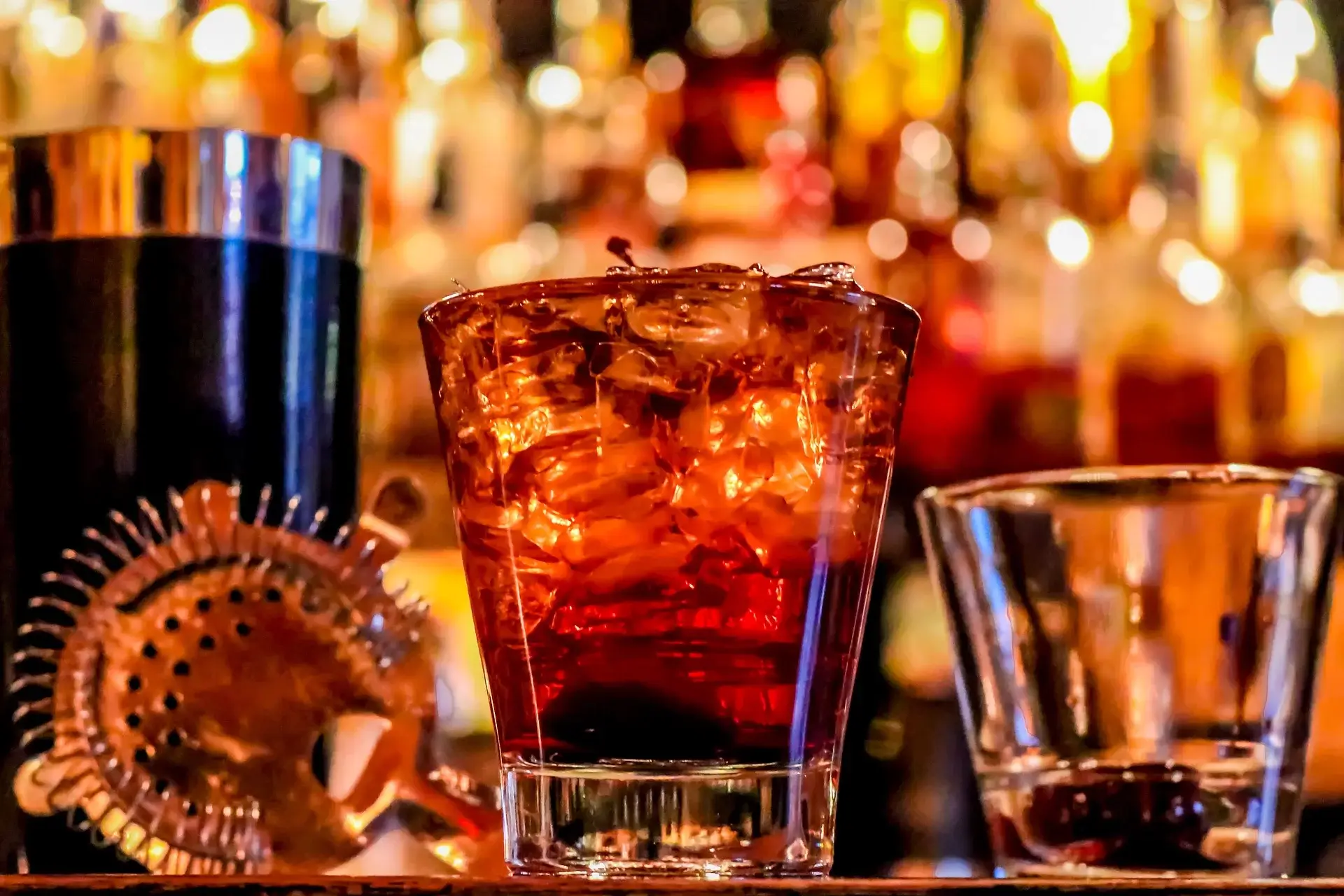 Glass of whiskey with ice cubes on a bar counter surrounded by a cocktail strainer and another empty glass.