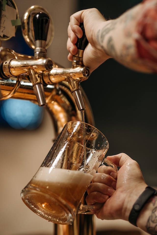 Hand with tattoos pulling a tap handle to pour beer into a tilted glass mug.