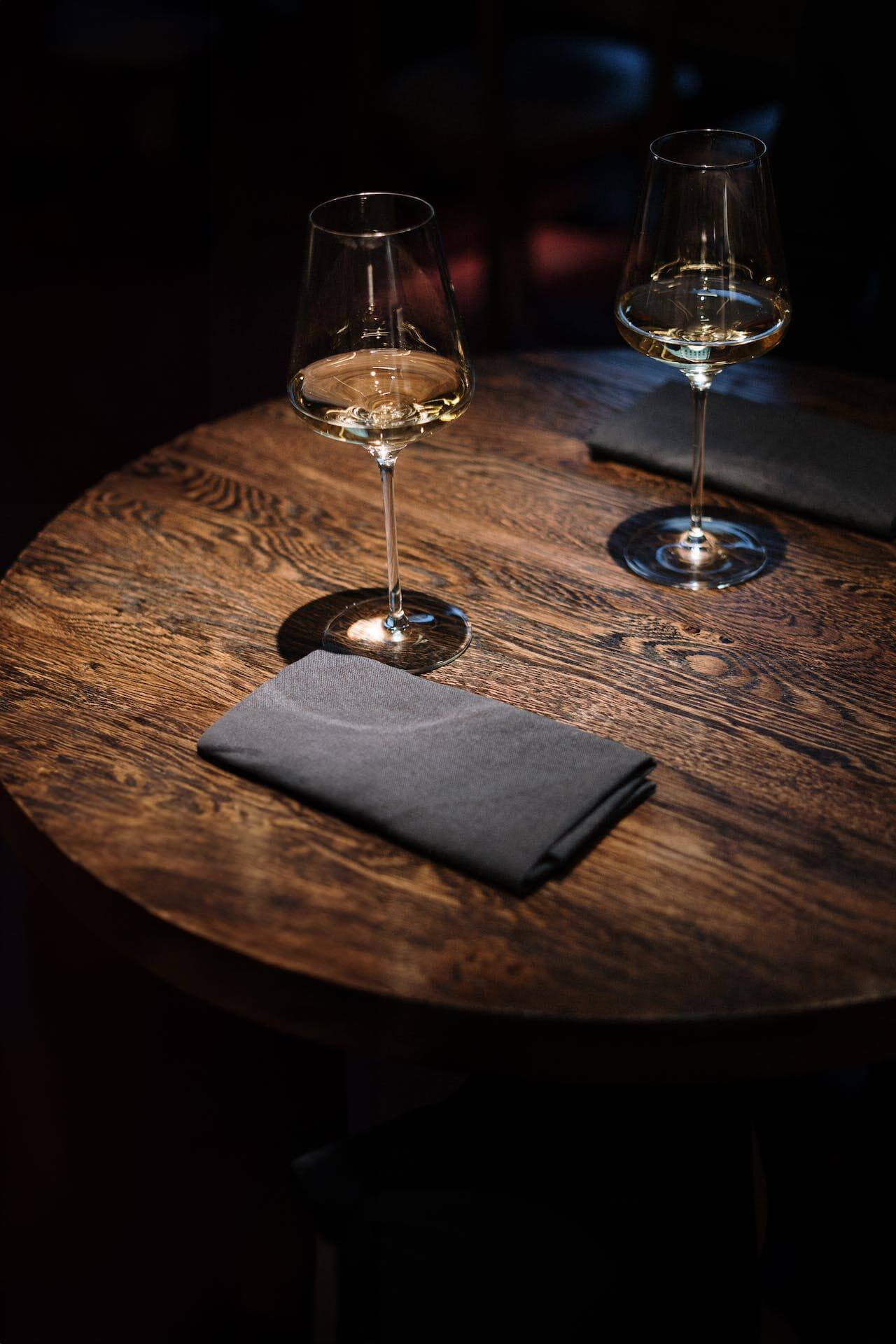 Two glasses of white wine and folded gray napkins on a round wooden table in dim lighting.