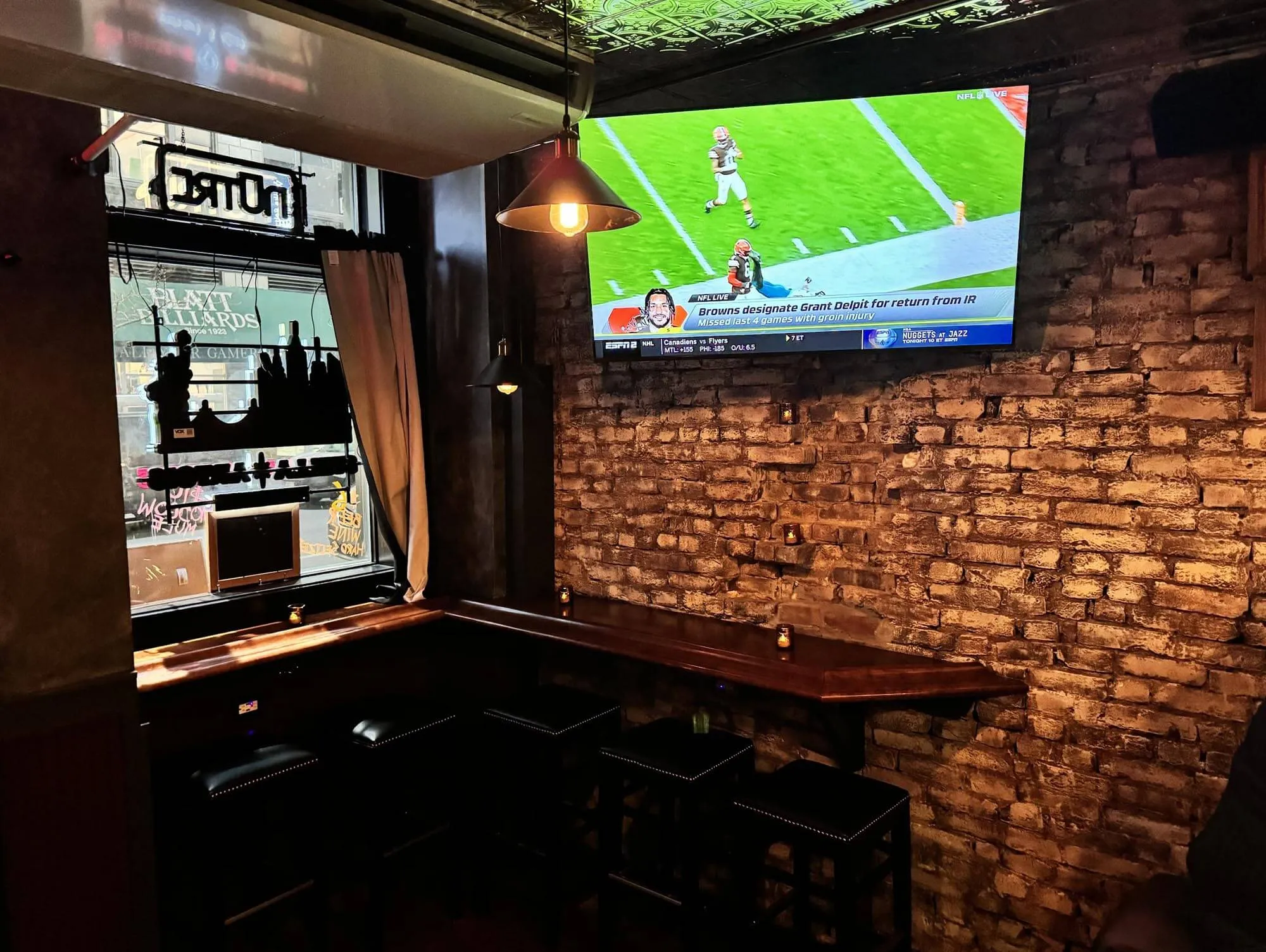 Rustic bar corner with exposed brick wall and wooden stools, a TV mounted on the wall showing an NFL game.